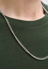 Una cadena de metal plateada se encuentra colgando sobre una camiseta de cuello redondo en color verde oliva, con una textura suave y eslabones uniformes.