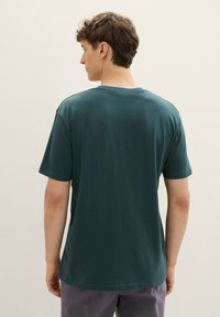 Camiseta de hombre de manga corta en algodón verde profundo. Cuello redondo sencillo, corte relajado, textura lisa. Sin logotipos ni patrones visibles. Vista trasera.