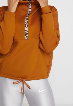 Personne ajustant le col d'un pull Nike Pro orange brûlé avec des leggings Nike blancs dotés d'une taille à cordon de serrage et d'un logo Swoosh.