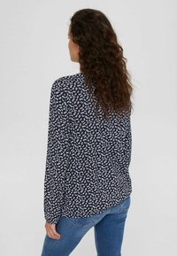 Donna con capelli lunghi e ricci, che indossa una blusa nera con una piccola stampa floreale bianca e jeans blu, in piedi di spalle contro uno sfondo neutro.