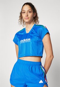 Zila īsā sporta krekls ar V veida kakla izgriezumu, Adidas logo, slaidām svītrām uz piedurknēm, apvienots ar atbilstošām zilām šortiem, kas piedāvā sānu svītras un logo.