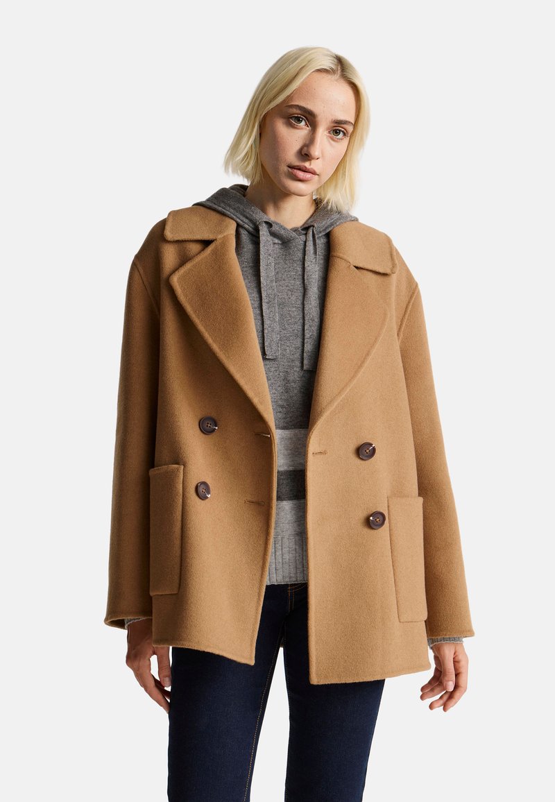 Kabát z camel vlny s dvojitým zapínáním, širokými klopami, dvěma předními kapsami a tmavě hnědými knoflíky, nošený přes šedou mikinu.