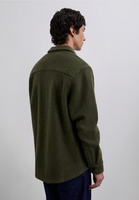 Chaqueta de mezcla de lana color verde oliva con puños abotonados, acabado texturizado y cuello clásico. Vista desde la parte trasera, mangas relajadas.