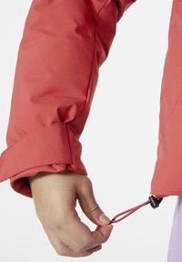 Roter, gepolsterter Jackenärmel mit elastischem Bündchen und verstellbarem Kordelzug. Besitzt einen schwarzen Toggle und einen glatten, wasserabweisenden Stoff.