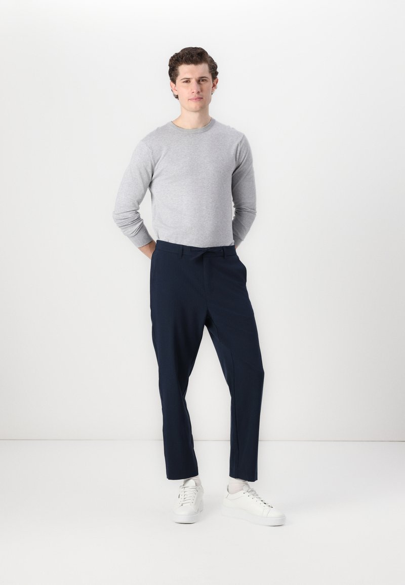 Un chandail gris clair à manches longues associé à un pantalon ajusté bleu marine et des baskets blanches. Les vêtements ont une texture lisse et une coupe moderne.