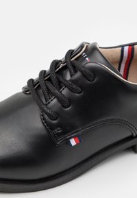 Czarne skórzane buty z okrągłym noskiem, czarnymi sznurowadłami i przeszyciem. Posiadają małą trójkolorową metkę z boku oraz gładką teksturę.