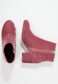 Par de botas de tobillo de terciopelo rosa con tacones de bloque y cremalleras laterales, mostradas desde arriba y de lado sobre un fondo blanco.