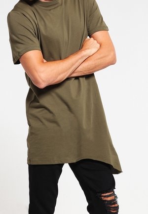 Personne en t-shirt oversize vert olive les bras croisés, portant un jean noir déchiré, sur un fond blanc uni.