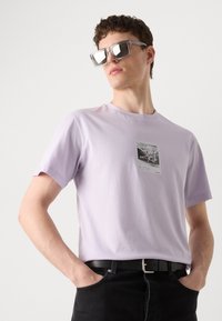 Lilac cotton t-shirt with a front graphic print in a rectangular frame. Model wears oversized gray sunglasses and black jeans with a belt.