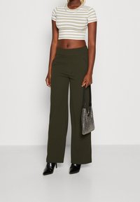 Gestreifter, kurz geschnittener Oberteil in Beige und Weiß, kombiniert mit hochgeschnittenen, olivgrünen, weit geschnittenen Hosen. Mit einer metallischen Handtasche accessoriert.