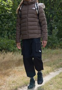 Bruine donsjas met een hoge kraag, zwarte cargobroek en zwarte wandelschoenen. Het terrein bestaat uit gras en een onverharde weg. Rugzak op de schouder.