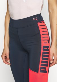 Leggings blu navy con accenti corallo presentano un grande logo nero "PUMA". Realizzati in tessuto elasticizzato, hanno una superficie liscia e un design aderente.