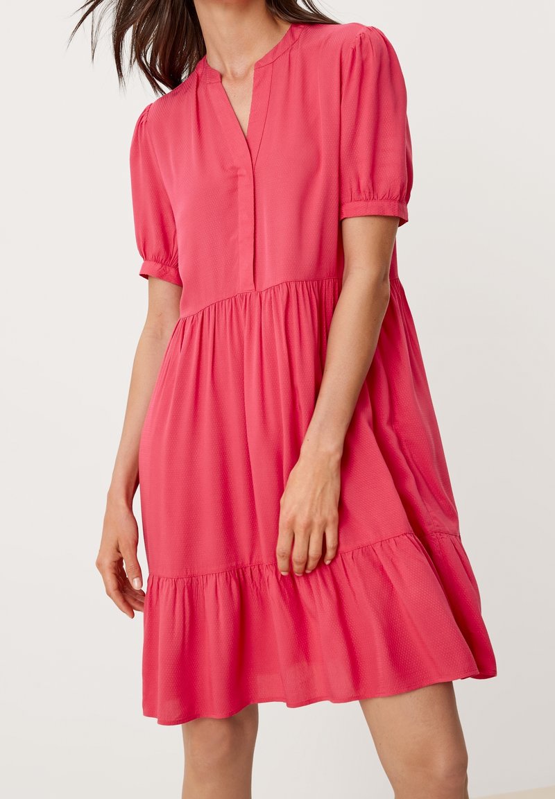 Femme portant une robe rose corail ample, à manches courtes, longueur genou, avec une jupe à volants superposés et un décolleté en V sur fond uni.