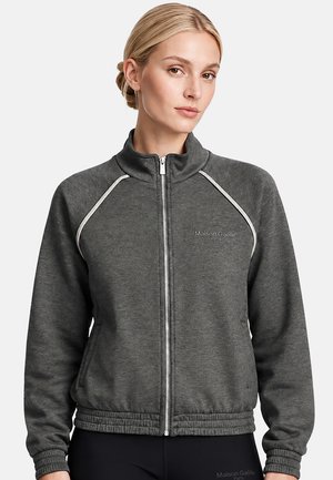Mujer con cabello rubio que lleva una chaqueta gris oscuro con cremallera, ribetes blancos y el logo "Maison Gaëlle Paris" en el pecho.
