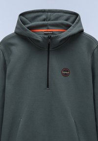 Mænds mørkegrøn hættetrøje med lynlås i halsen, med en orange inderside og et sort cirkulært logo på brystet.