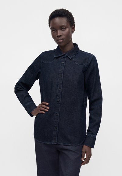 Personne à la peau foncée portant une chemise en denim bleu foncé à boutons avec col, debout avec une main sur la hanche sur un fond uni.
