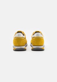 Le scarpe da corsa gialle in suede presentano una intersuola grigia e una suola in gomma, con un accentuazione del tallone bianca e un logo sul retro.