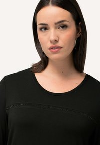Schwarzes Langarmshirt aus weichem Material mit rundem Ausschnitt, das entlang der Brust eine horizontale Linie kleiner dekorativer Akzente aufweist.