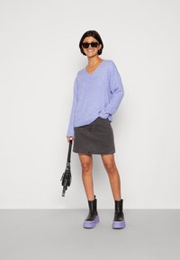 Pull léger violet en V, jupe en denim gris foncé, sac à main noir et bottes plateforme noires et violettes avec une semelle épaisse. Textures de tissu lisses visibles.