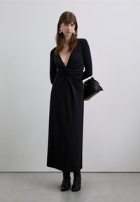 Robe longue noire avec un profond décolleté en V et un détail torsadé au centre. Fabriquée en tissu doux, associée à des bottines à talons noires élégantes et une petite pochette noire.