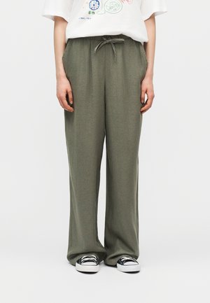 Person trägt lockere olivgrüne Hosen mit Kordelzug, weißes T-Shirt mit Grafik und schwarz-weiße Turnschuhe und steht vor einem schlichten Hintergrund.