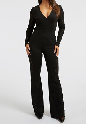 Kobieta ubrana w dopasowany czarny kombinezon z koronkowymi długimi rękawami i koronkowymi panelami wzdłuż szerokich spodni, w połączeniu z czarnymi szpiczastymi czółenkami.