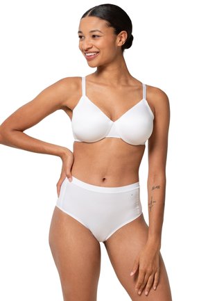 Femme souriante avec les cheveux attachés, portant un soutien-gorge blanc et une culotte taille haute, debout avec une main sur la hanche et l'autre détendue.