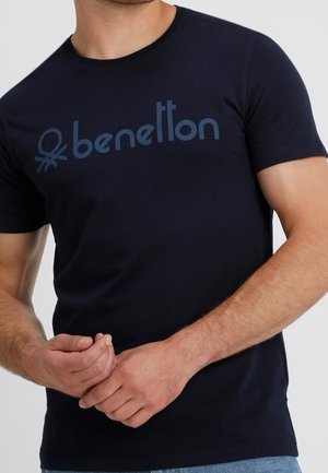 Mężczyzna w granatowej koszulce z krótkim rękawem z logo "benetton" w kolorze niebieskim na piersi, stoi ze splecionymi dłońmi przed sobą.
