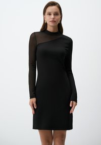Zwarte jurk met lange, doorzichtige mesh-mouwen en een hoge hals. Vloeiende stof, getailleerde vorm en asymmetrische halslijn. Eenvoudig ontwerp.