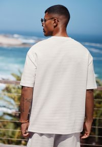 Vit texturerad kortärmad skjorta på en person, sedd bakifrån, med rund halsringning och avslappnad passform. Havet syns i bakgrunden.