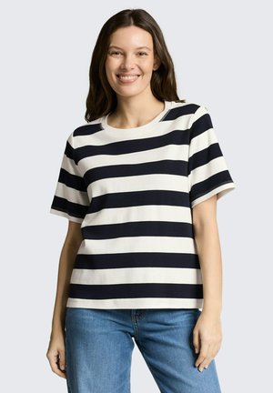 Frau lächelt, trägt ein schwarz-weiß gestreiftes T-Shirt mit kurzen Ärmeln und blaue Jeans, steht vor einem hellgrauen Hintergrund.