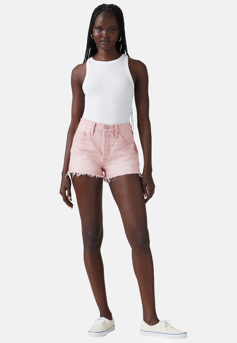 Lichtroze denimshorts met een rafelige zoom, hoge taille; gecombineerd met een witte, strakke tanktop en lichtgekleurde sneakers.