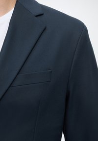 Gros plan sur l'épaule et la poche poitrine d'un blazer bleu marine foncé porté sur une chemise blanche sur une personne.