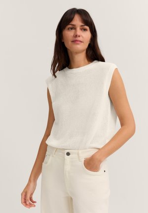 Frau mit langen dunklen Haaren, die ein weißes ärmelloses Stricktop und weiße Hose trägt, steht mit einer Hand in der Tasche vor einem schlichten Hintergrund.