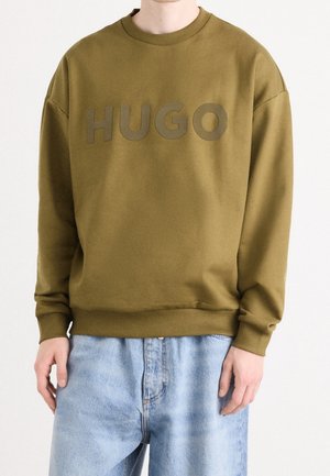 Osoba v olivově zelené mikině s vyvýšeným logem "HUGO" na hrudi a světle modrých džínách, stojící před bílým pozadím.