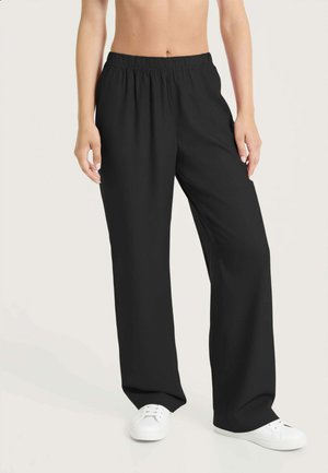 Persona che indossa pantaloni neri larghi con vita elastica e scarpe da ginnastica bianche, in piedi davanti a uno sfondo chiaro e uniforme.