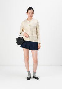 Kvinne i en kremfarget kabelstrikket cardigan, marineblå plisserte skjørt, grå sokker, svarte ballettsko, og holder en liten svart håndveske.