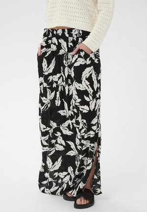 Czarna maxi spódnica z białym nadrukiem liści, bocznym rozporkiem, elastycznym pasem, noszona z białym swetrem dzianinowym i czarnymi sandałami. Ręce w kieszeniach.