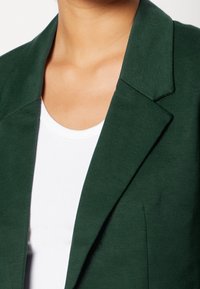 Tmavě zelený blazer se strukturovaným límcem a klopami, s hladkým povrchem. Nosený přes bílé tričko s širokým výstřihem.