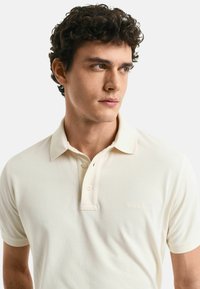 Ljusbeige polo skjorta i mjukt tyg, med spetsig krage, korta ärmar och en subtil logotyp på bröstet.