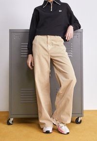 Maglione nero con zip quarti e bordi bianchi, pantaloni beige a gamba larga e scarpe da ginnastica multicolori in piedi davanti a un armadietto grigio.