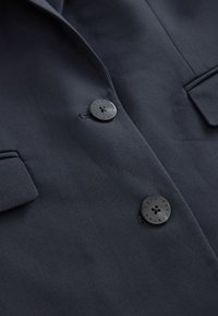 Blazer bleu marine foncé avec deux boutons noirs marqués "Next Tailoring", avec un revers cranté et une poche à rabat.