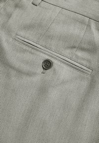 Close-up of beige textured fabric featuring a buttoned welt pocket with four-hole dark button sewn below the pocket slit.