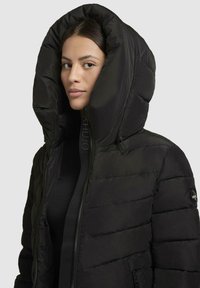 Black puffer jacket with a large hood, zip closure, and quilted texture. Features branded hardware and side pockets. Smooth, glossy finish.