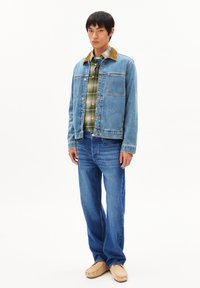 Denimjack met een bruine corduroy kraag, twee borstzakken, een blauw geruit shirt eronder, gecombineerd met blauwe jeans en beige schoenen.