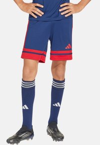 Blaue Sportshorts mit roten Streifen am Saum, kombiniert mit blauen kniehohen Socken mit weißen Streifen und schwarzen Fußballschuhen.