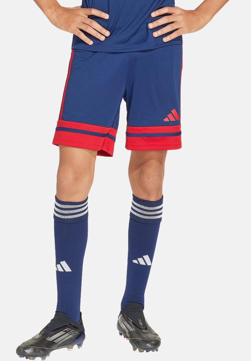 Blaue Sportshorts mit roten Streifen am Saum, kombiniert mit blauen kniehohen Socken mit weißen Streifen und schwarzen Fußballschuhen.