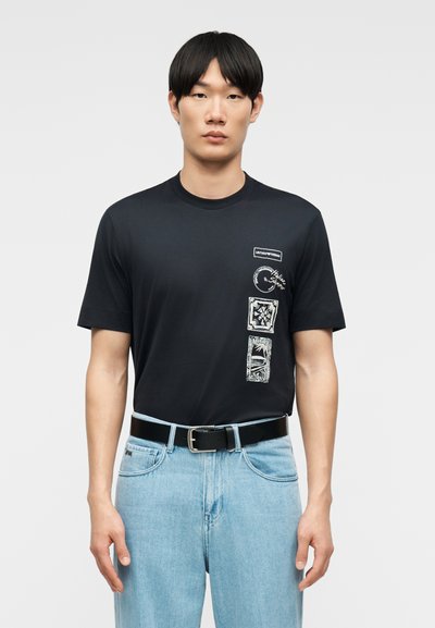 Άνδρας που φοράει μαύρο γραφικό t-shirt με λευκά σχέδια στο αριστερό στήθος, φωτεινό μπλε τζιν και μαύρη ζώνη, στέκεται μπροστά σε απλό γκρι φόντο.