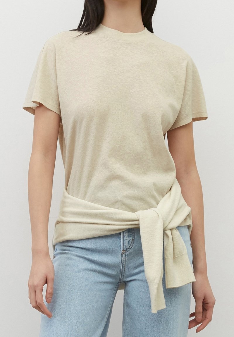 Femme portant un haut beige à manches courtes avec un pull assorti noué autour de la taille et un jean bleu clair, sur un arrière-plan uni.