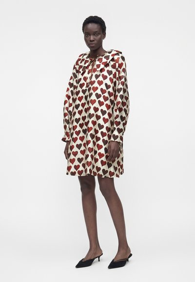 Γυναίκα που φοράει ένα άσπρο φόρεμα μέχρι τα γόνατα με κόκκινα και καφέ σχέδια σε σχήμα καρδιάς, συνδυασμένο με μαύρα μυτερά τακούνια.